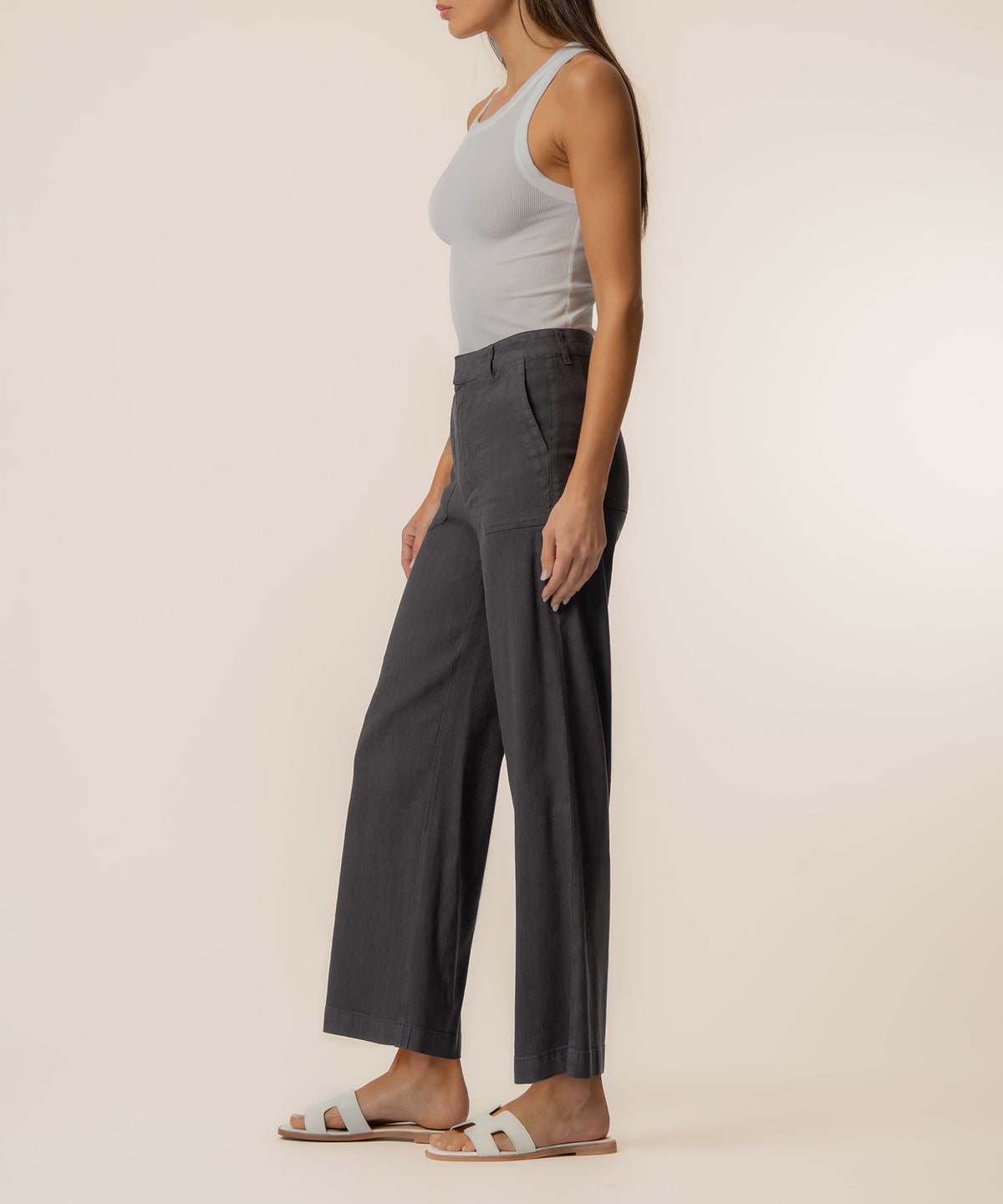 Woman wearing charcoal wide-leg linen pants with white tank top and white slide sandals, shown from the side.