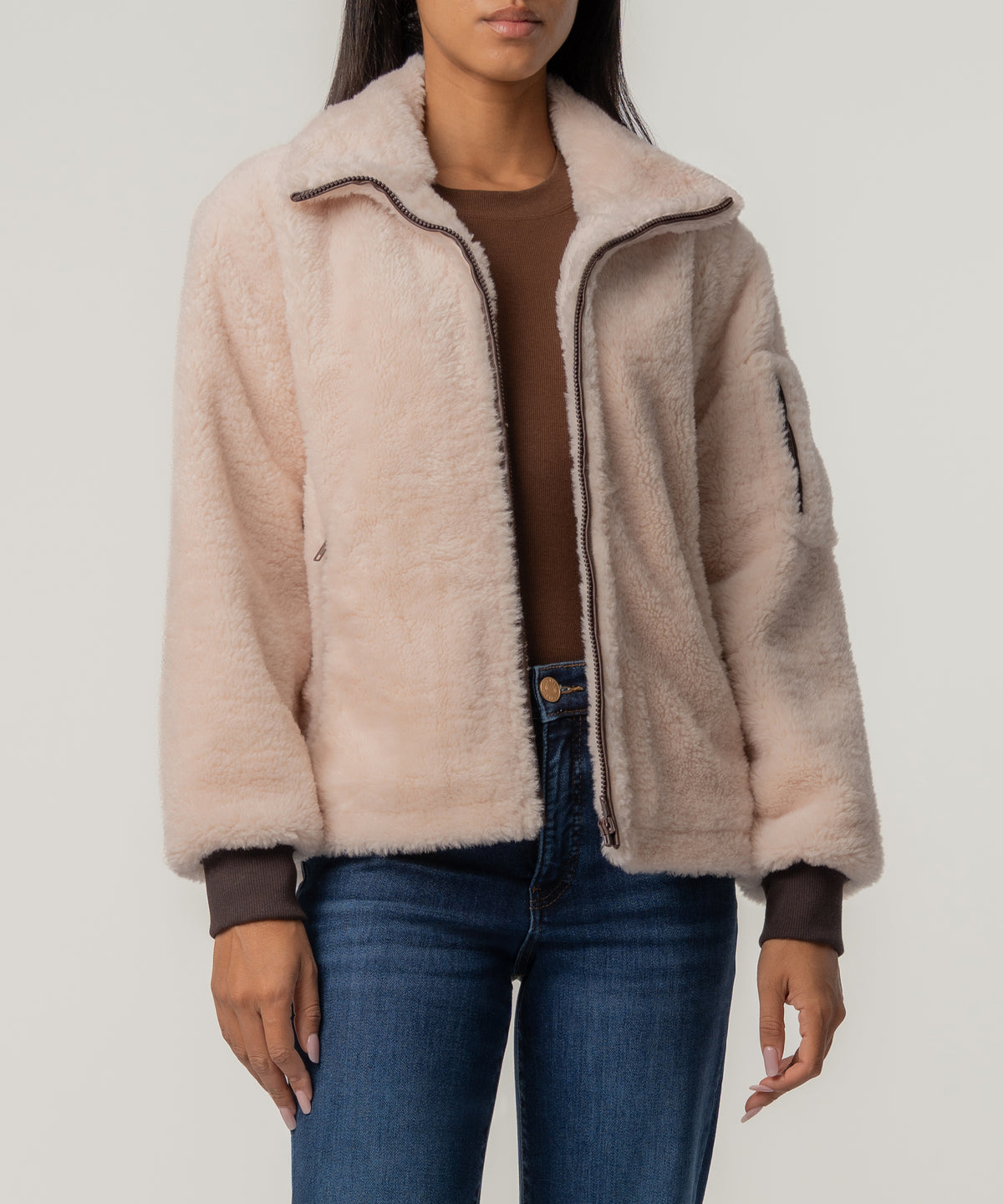 A woman wears a cream faux sherpa jacket with brown zipper and ribbed cuffs over a brown top and blue jeans.