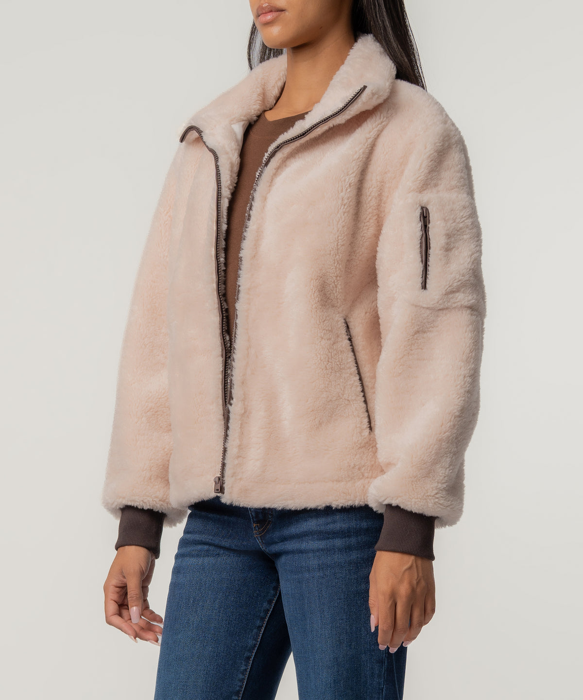 A woman wears a cream faux sherpa jacket with a brown zipper, ribbed brown cuffs, a sleeve pocket, and blue jeans.