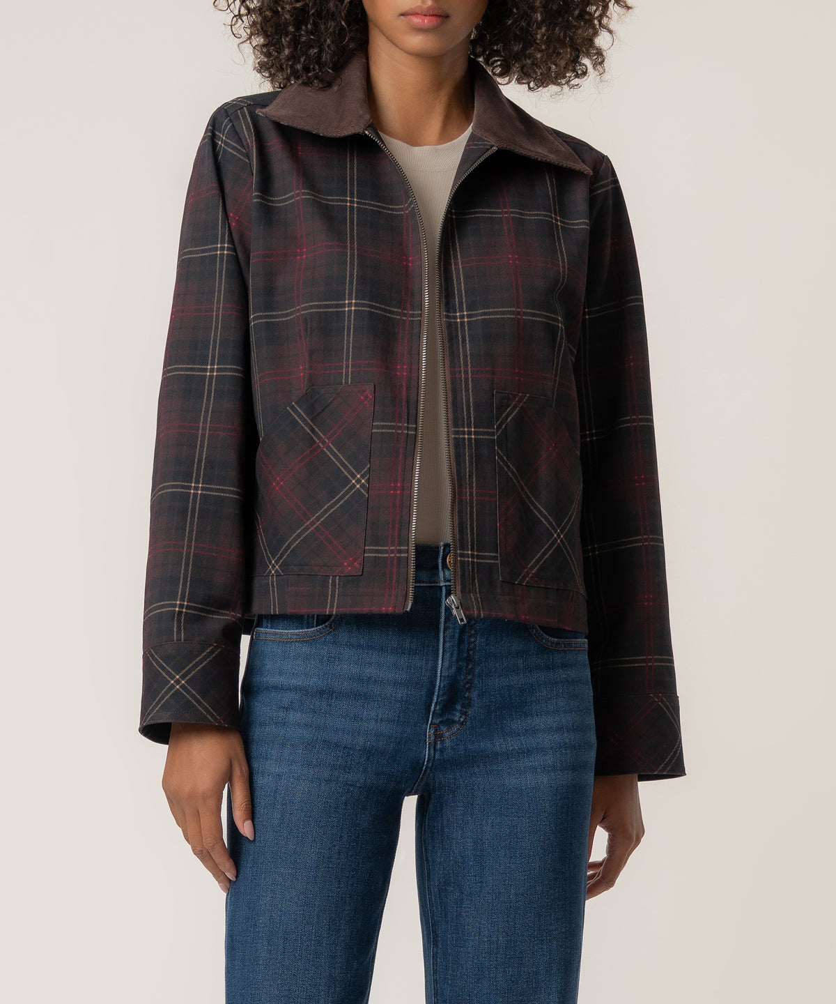 A woman wears a dark plaid zip-front jacket with a brown corduroy collar and patch pockets over blue jeans.
