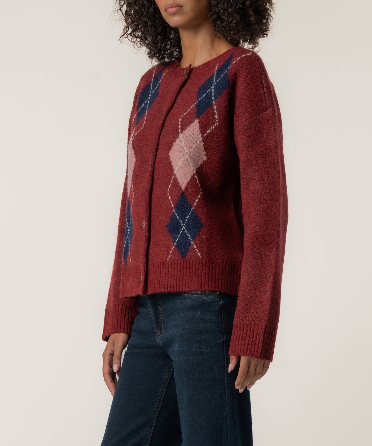 A woman with curly hair wears a dark red button-front cardigan with navy and pink argyle diamonds, paired with dark blue jeans.