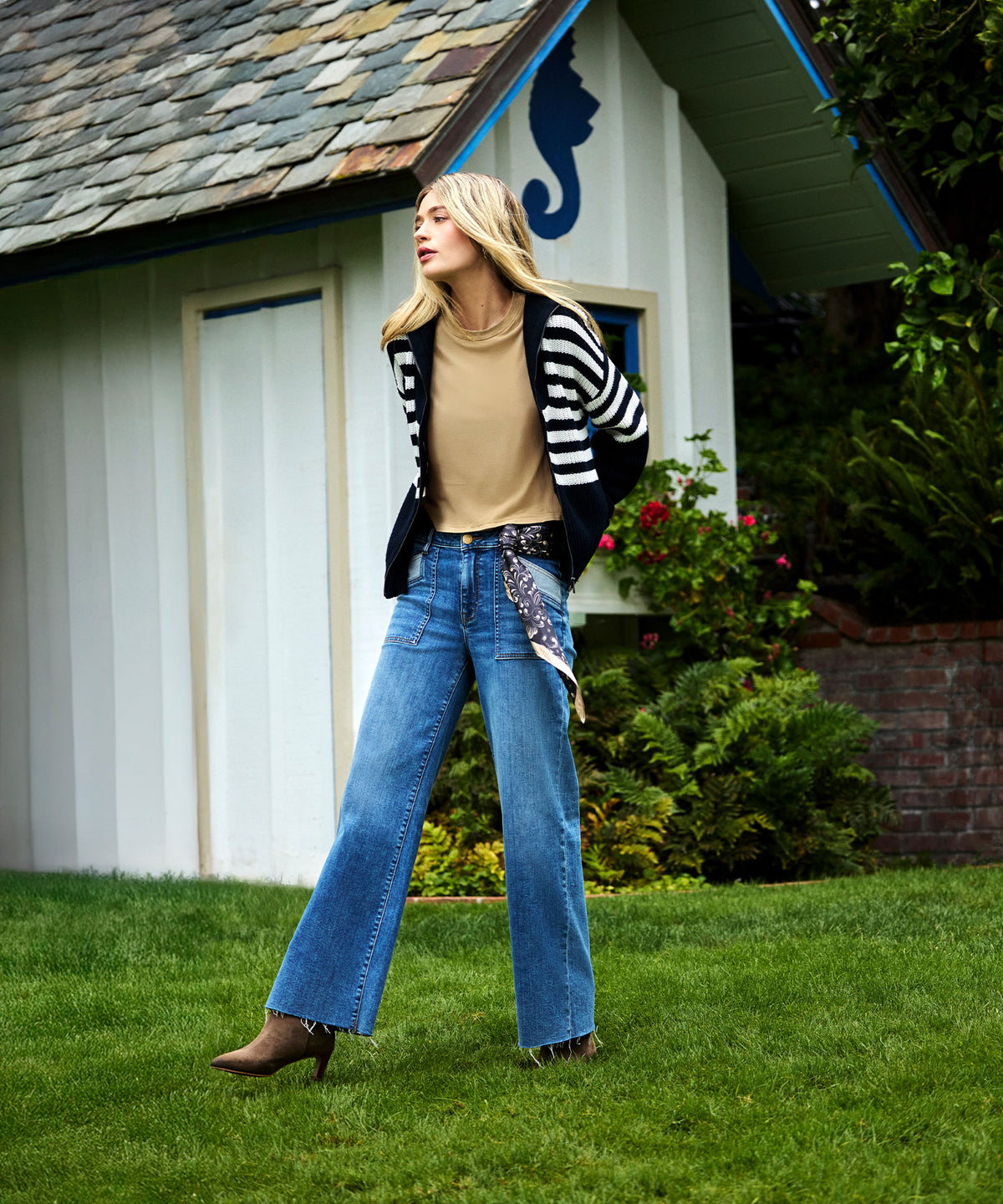 Woman wearing light blue high-rise wide-leg jeans with a cream top and striped cardigan, standing on grass in front of a hous