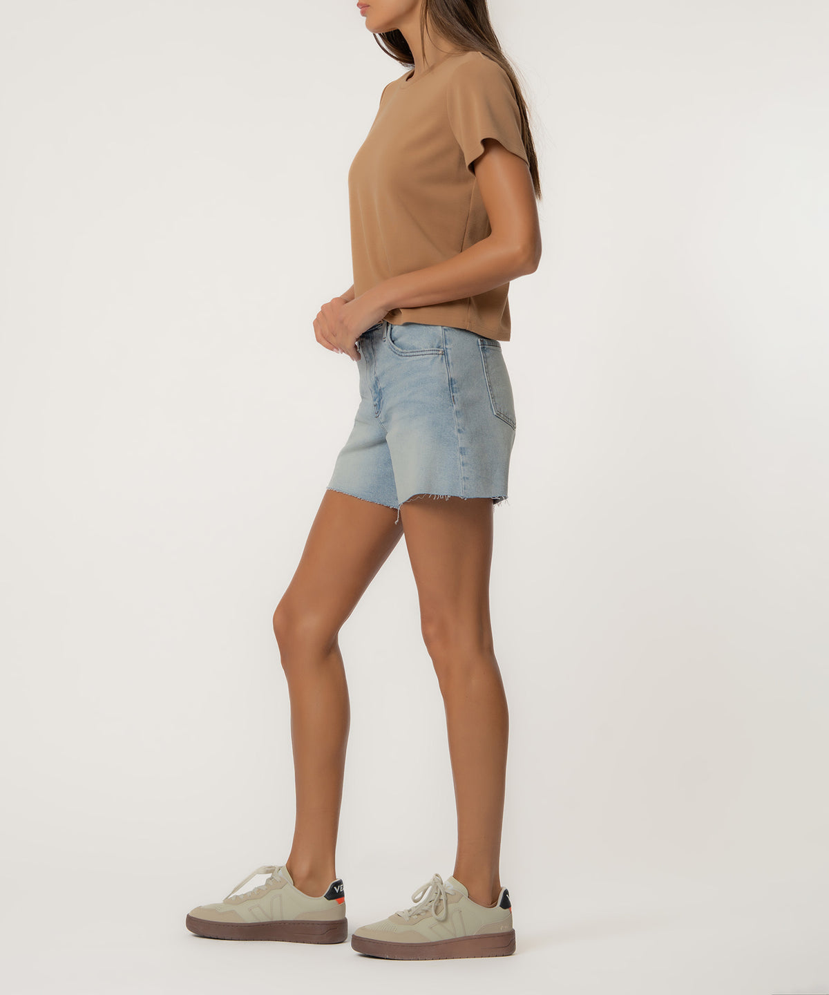 Woman wearing light wash denim shorts with high rise waistband and raw-cut hem, paired with tan t-shirt.