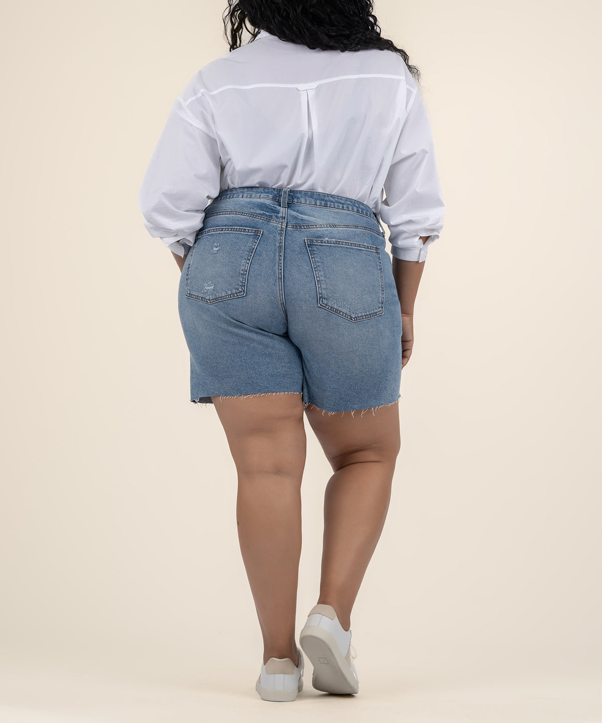 Rear view of a person wearing light blue denim Bermuda shorts with frayed hems, a white button-up shirt, and white sneakers.