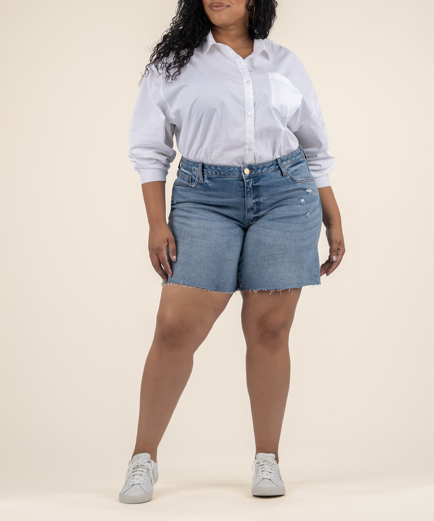 A woman wears medium-wash denim Bermuda shorts with a raw frayed hem, a white button-up shirt, and white sneakers.