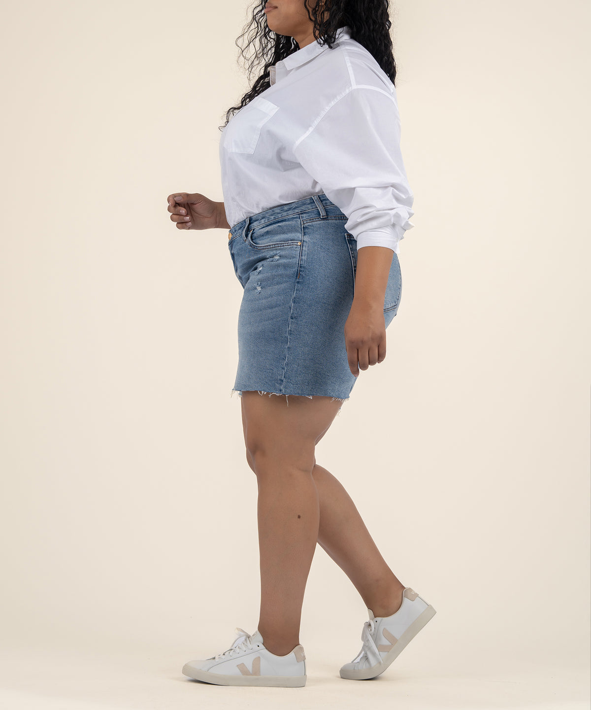 A woman in profile wears light blue denim shorts with a frayed hem, a white button-up shirt, and white sneakers.