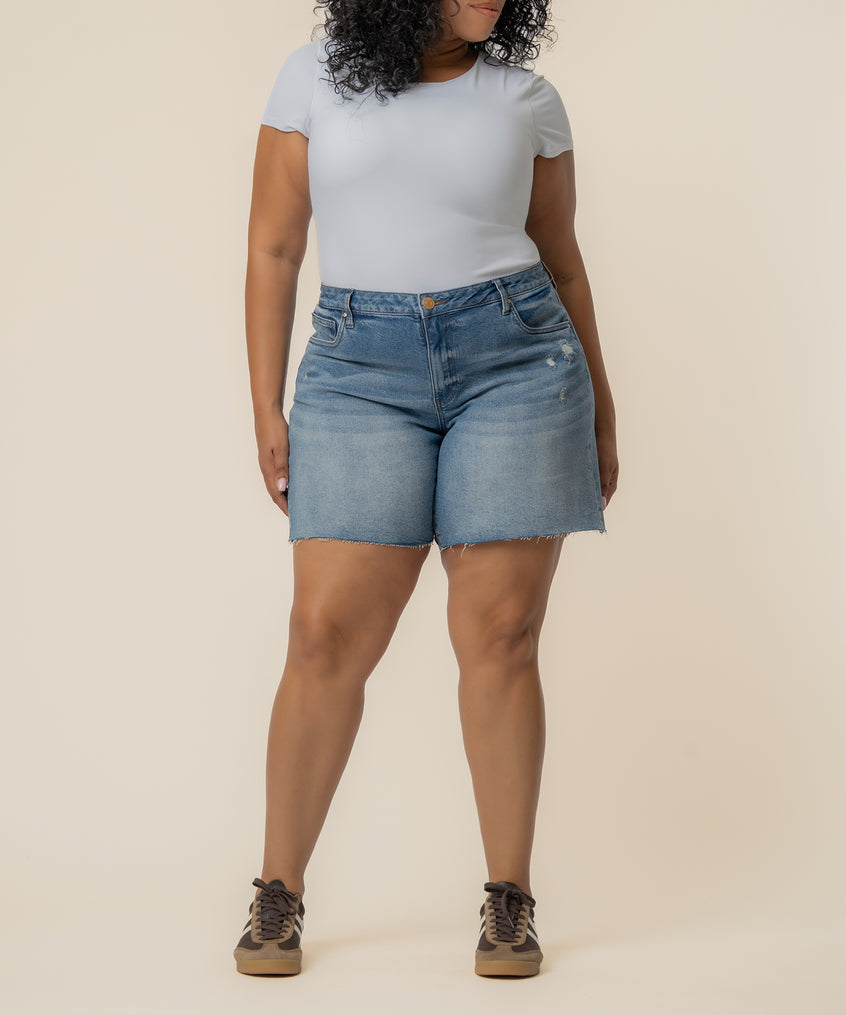 Woman wearing light blue denim bermuda shorts with high rise waist and distressed hem, paired with white t-shirt.