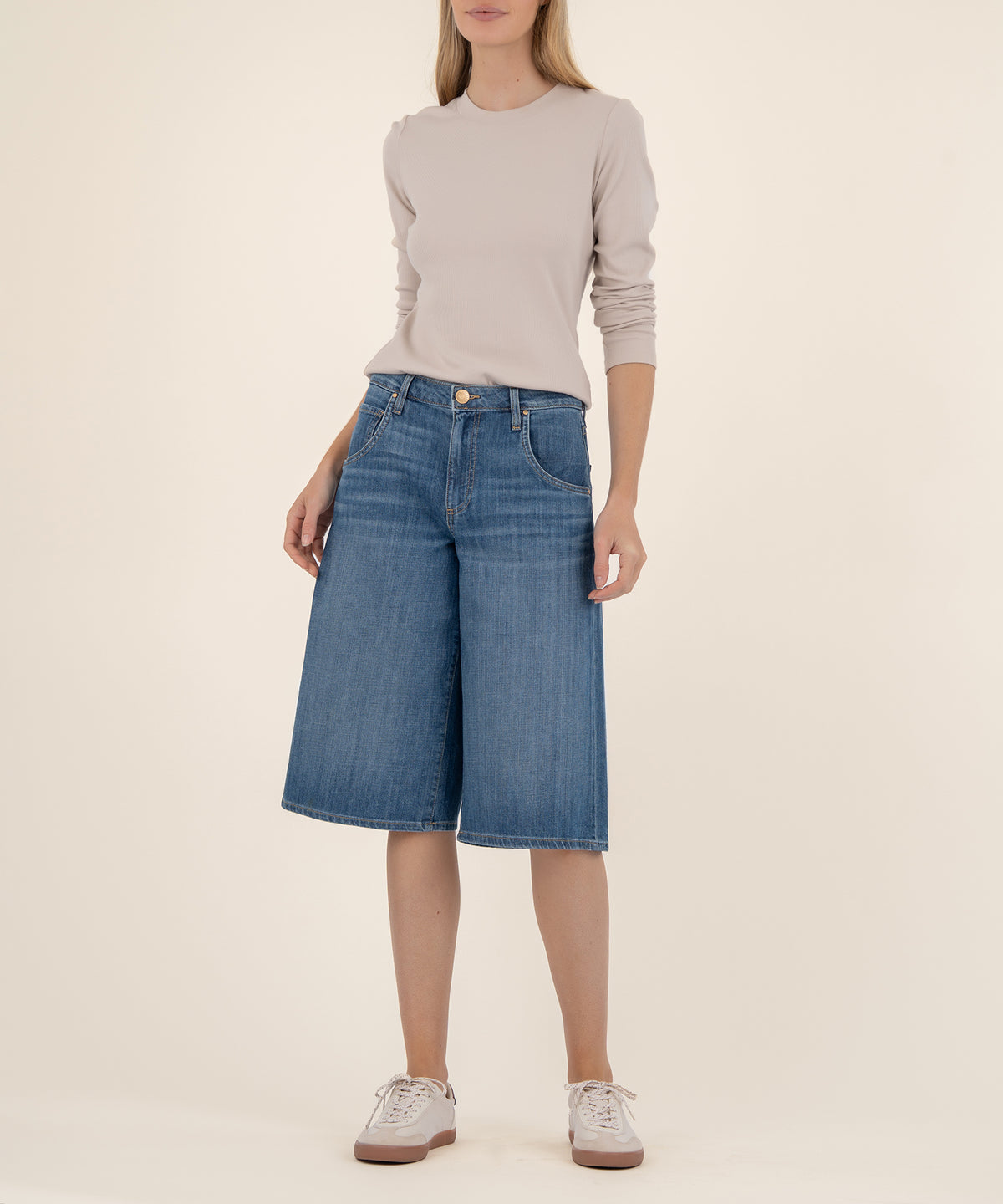 Woman wearing beige long-sleeve top and blue denim high-rise bermuda shorts with white sneakers.