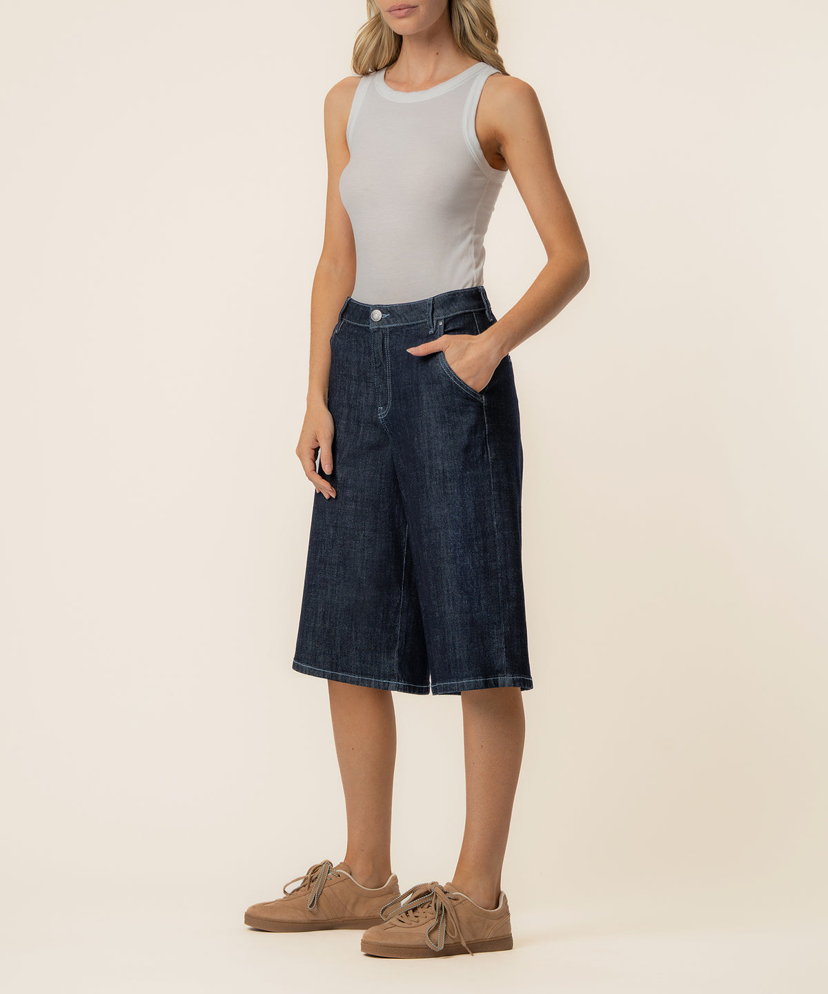 Woman wearing light gray sleeveless tank top and dark indigo denim bermuda shorts with front pockets.