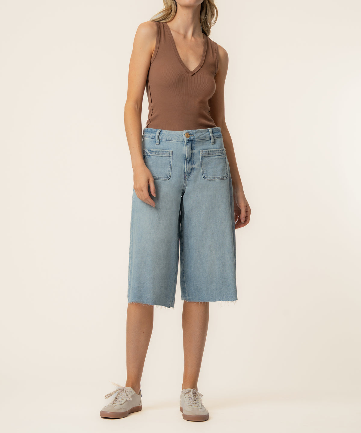 Woman wearing light blue denim high-rise bermuda shorts with raw hem and brown sleeveless tank top.
