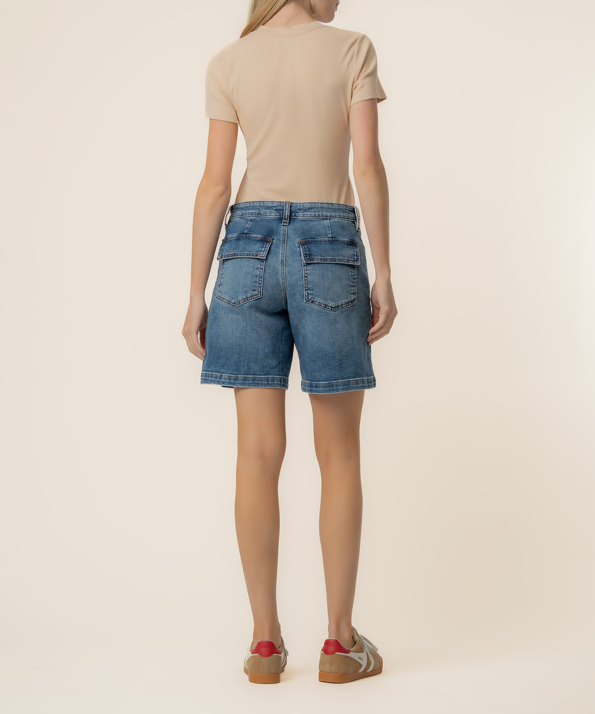 Woman wearing a beige t-shirt and blue denim high-rise bermuda shorts with patch pockets, viewed from behind.