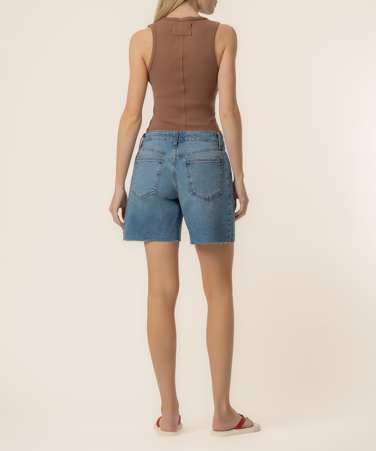 Woman wearing light blue denim bermuda shorts with raw hem and brown sleeveless top, viewed from back.