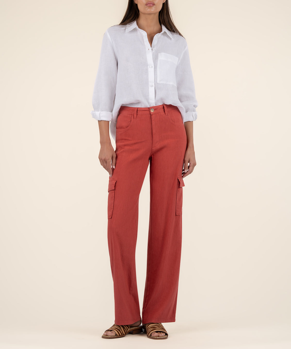 A woman stands against a cream background wearing rust-red wide leg cargo pants, a white linen shirt, and striped sandals.