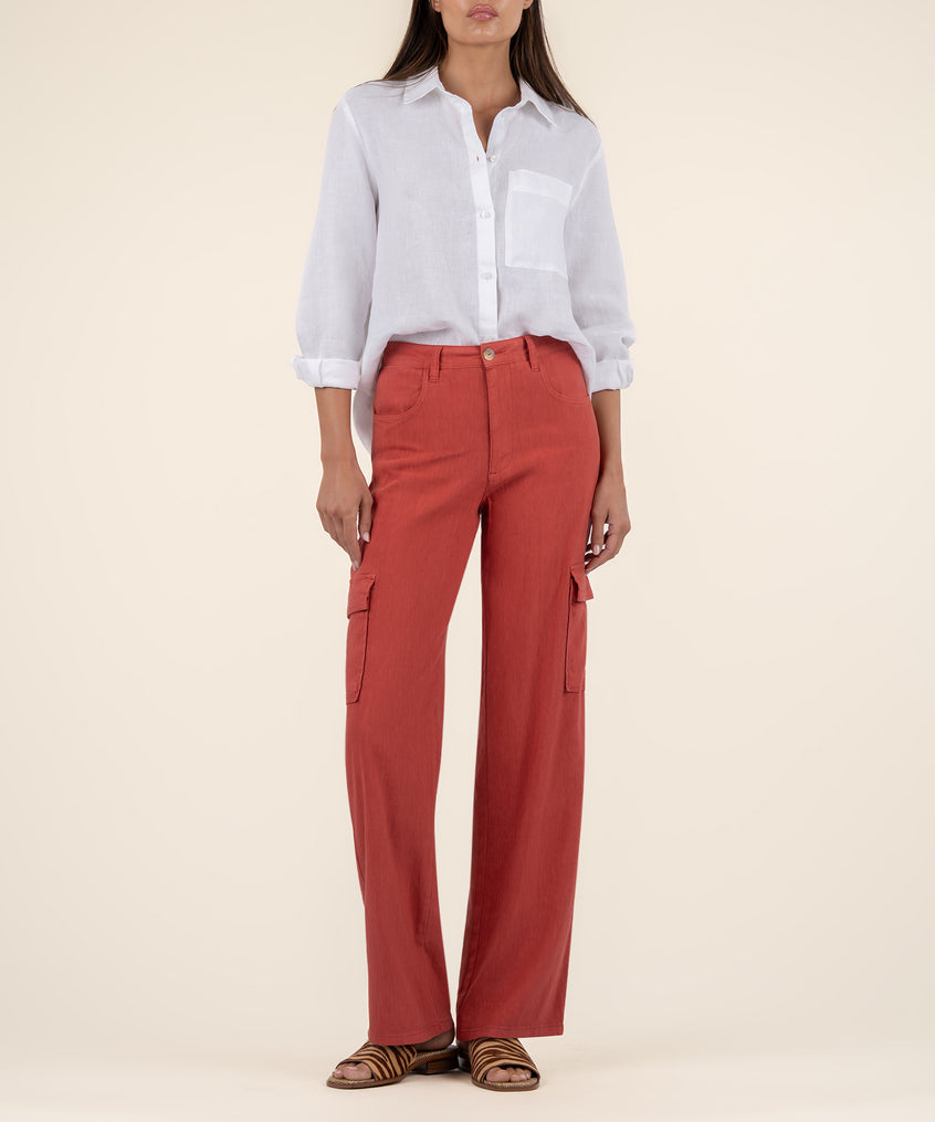 A woman stands against a cream background wearing rust-red wide leg cargo pants, a white linen shirt, and striped sandals.
