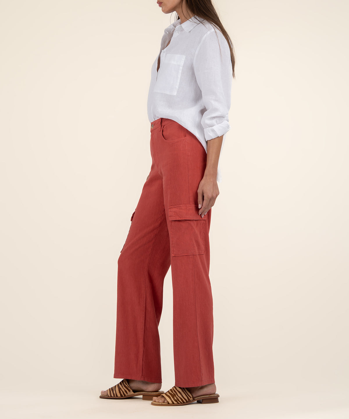 A woman stands in profile wearing rust-red wide leg cargo pants, a white linen shirt, and animal-print sandals.