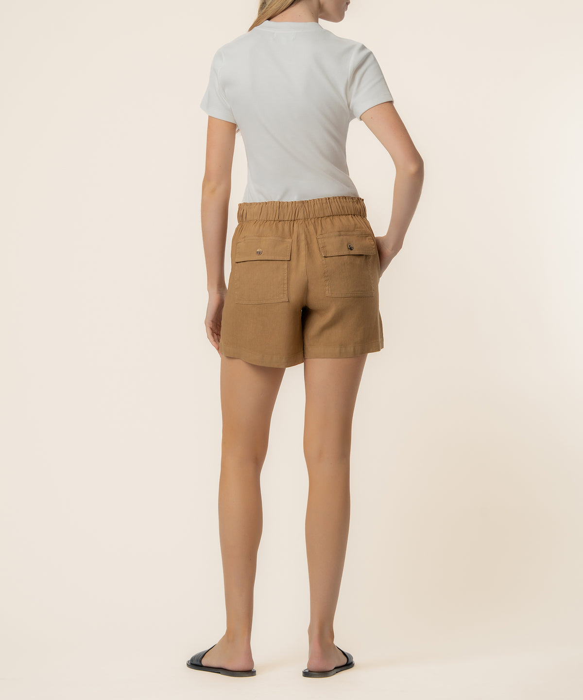 Woman wearing tan linen shorts with drawstring waist and back patch pockets, paired with white t-shirt.