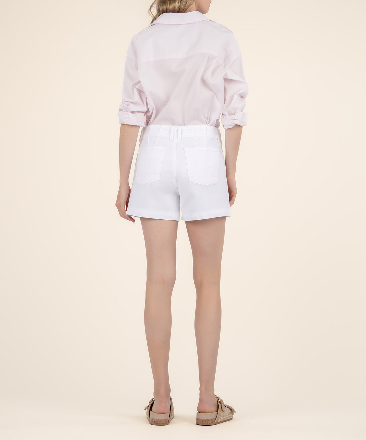 Rear view of a model wearing white high-rise shorts with two back pockets, a light pink shirt, and tan sandals.