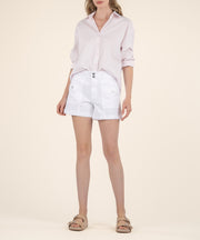 A woman wears white high-rise shorts with two front buttons and patch pockets, a light pink button-up shirt, and tan sandals.
