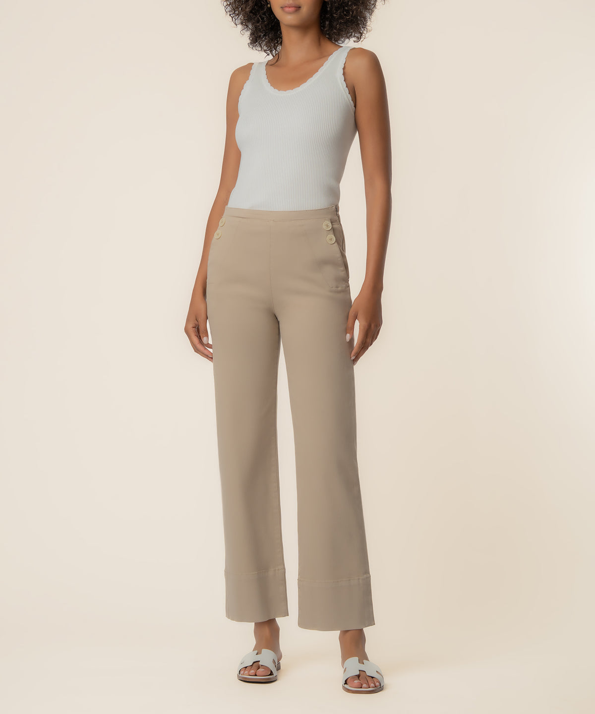 Woman wearing sand-colored high-rise wide leg twill pants with button details at waist, paired with white ribbed tank top.