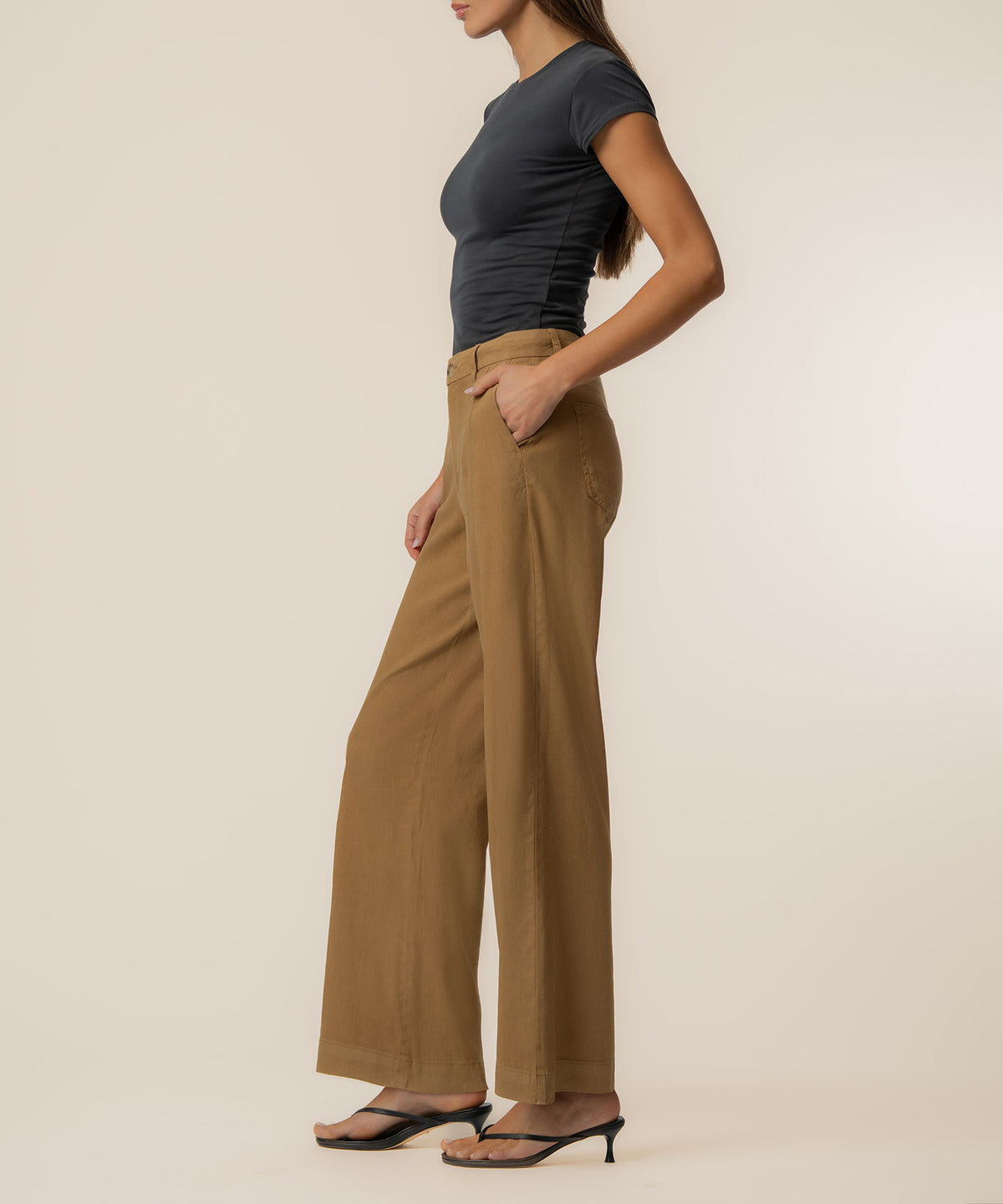 Woman wearing tan linen high-rise wide-leg pants with black short-sleeve top and black strappy heels, shown in side profile.