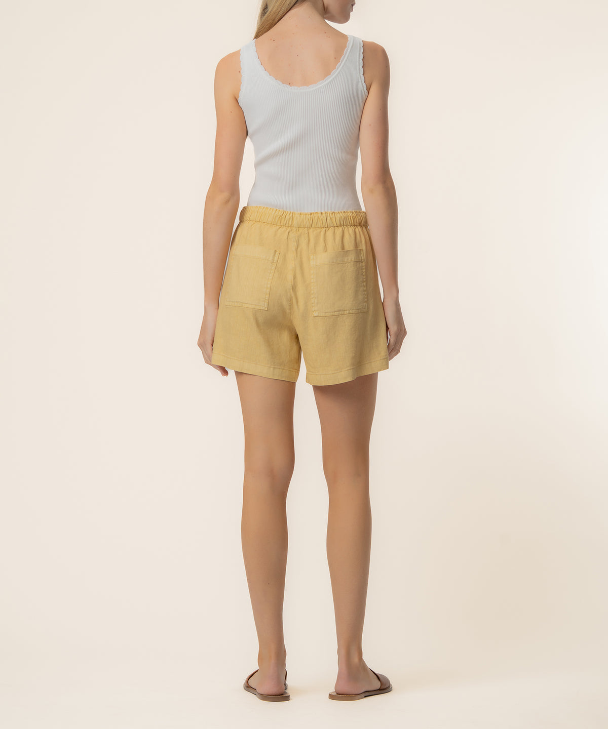 Woman wearing mustard gold linen shorts with high rise waistband and back patch pockets, paired with white ribbed tank top.