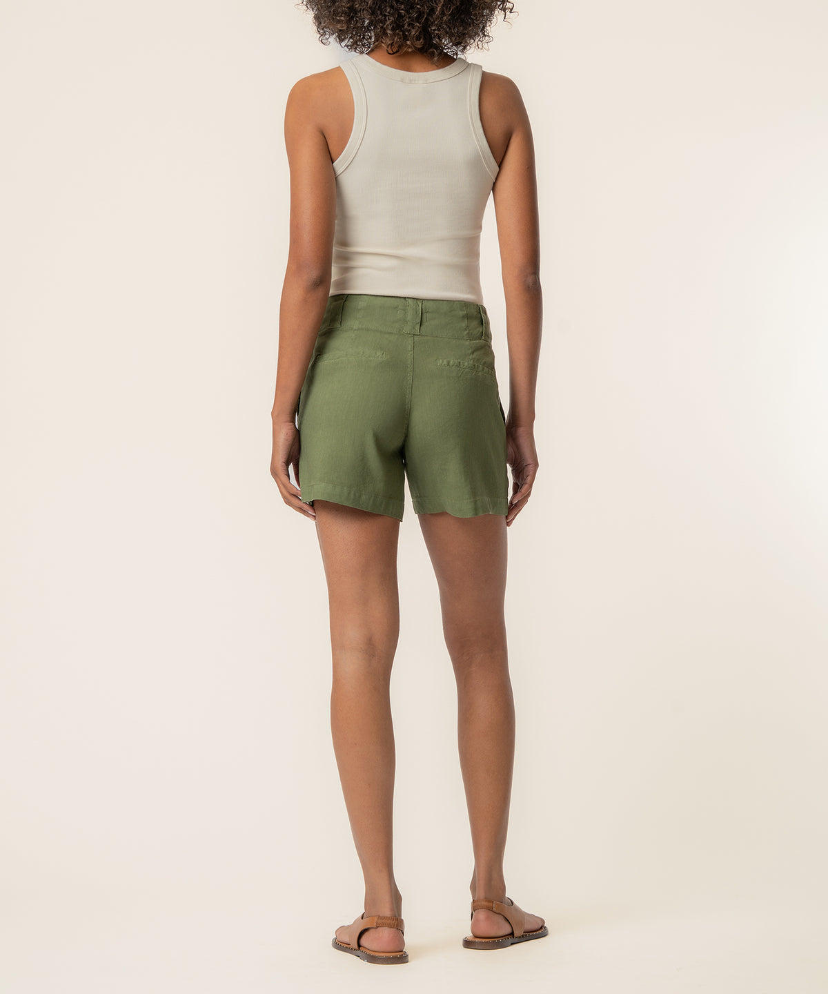 Woman wearing a sage green high-rise linen short with a white tank top, shown from behind.