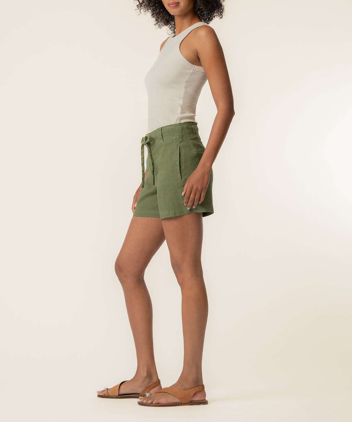 Woman wearing a dill-green linen high-rise short with a white tank top and brown sandals.