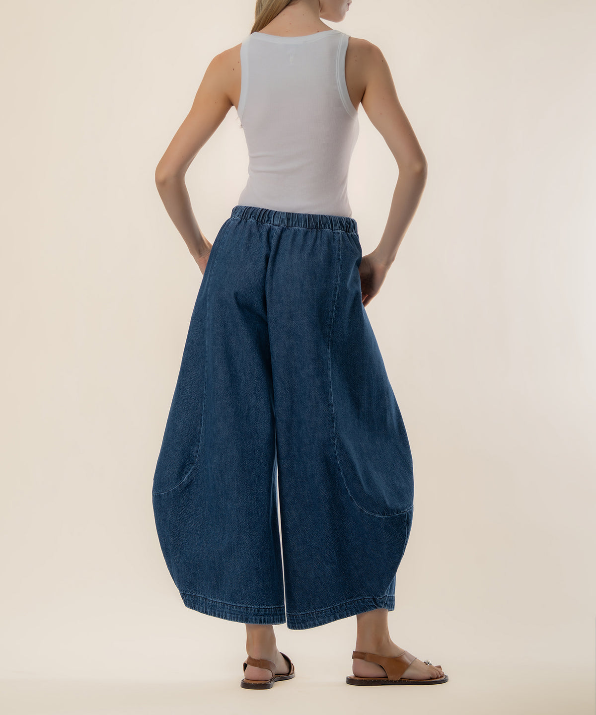 Woman wearing white sleeveless tank top and dark blue denim baggy barrel jeans with elastic waistband, viewed from behind.