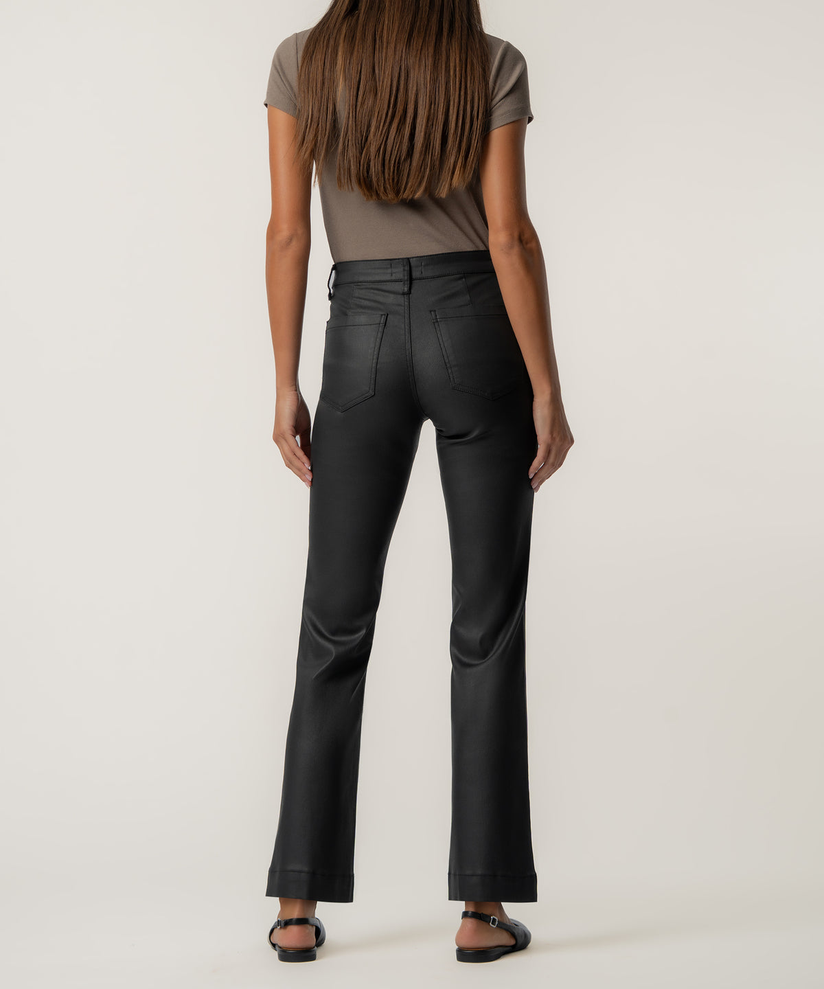 Back view of a woman wearing high-rise black coated flare pants with back pockets, a taupe short-sleeve top, and black flat sandals.