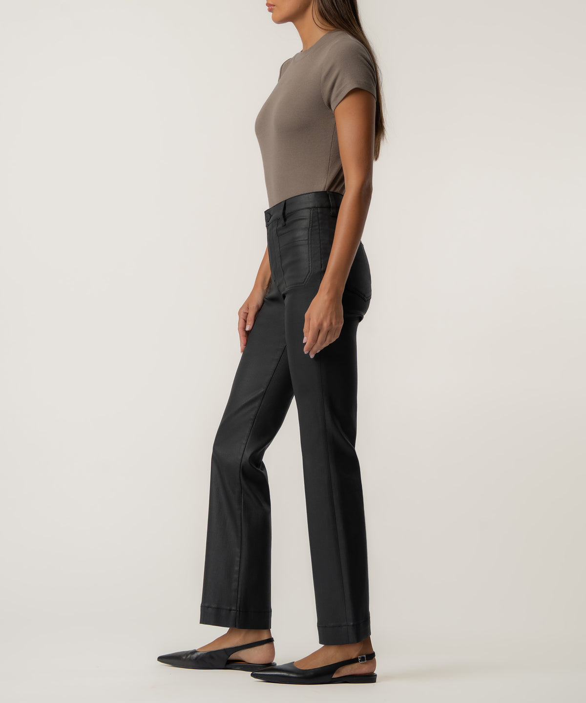 A woman stands in profile wearing high-rise black coated flare pants, a taupe short-sleeve top, and black pointed slingback flats.