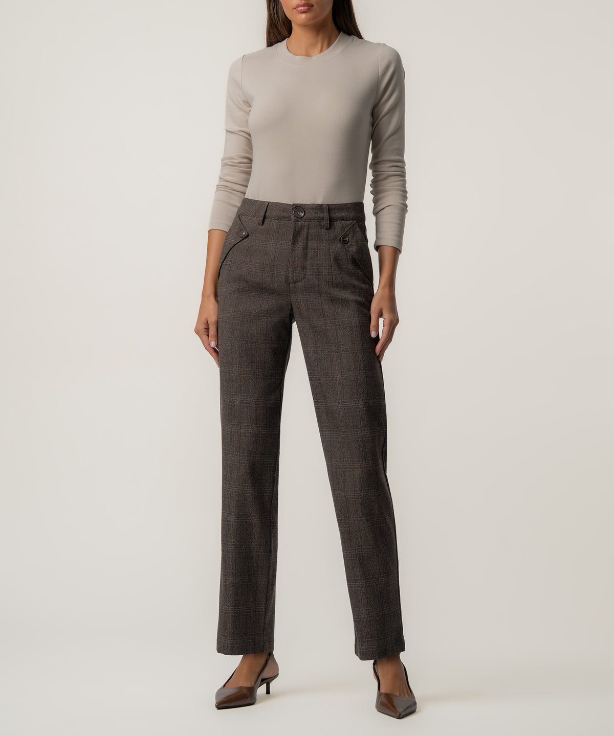 A woman wears high-rise dark brown plaid trousers with a beige long-sleeve top and brown pointed kitten heels.