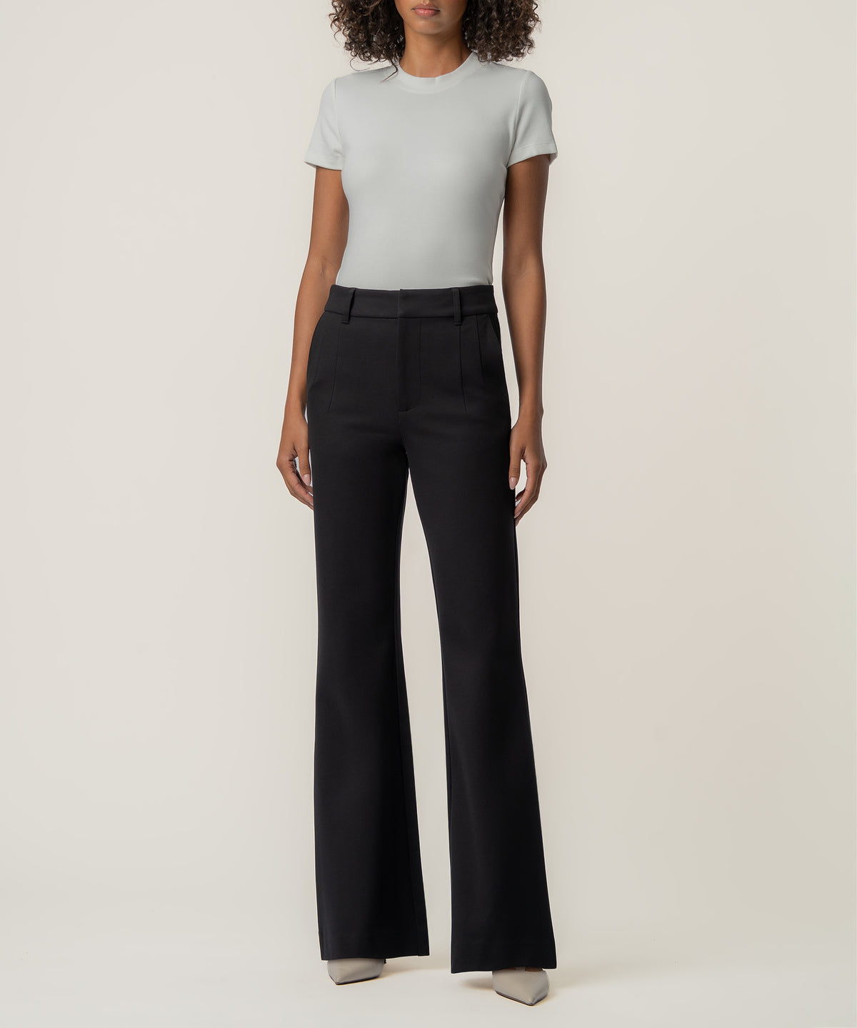A woman stands facing forward in high-rise black flare pants, a fitted light gray short-sleeve top, and pointed beige shoes.