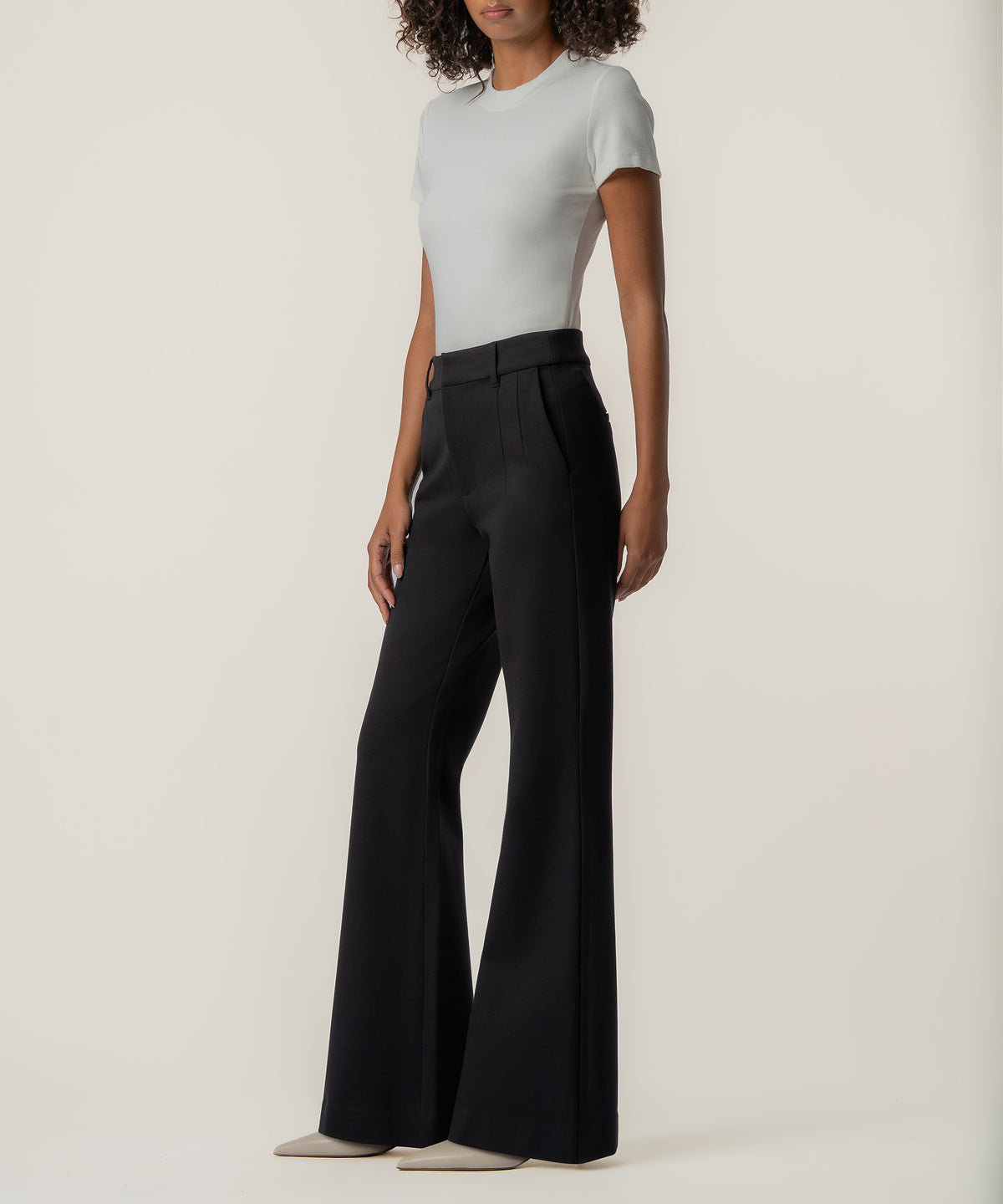 A woman wearing high-rise black flare pants with a fitted pale gray short-sleeve top and beige pointed-toe shoes stands against a neutral background.