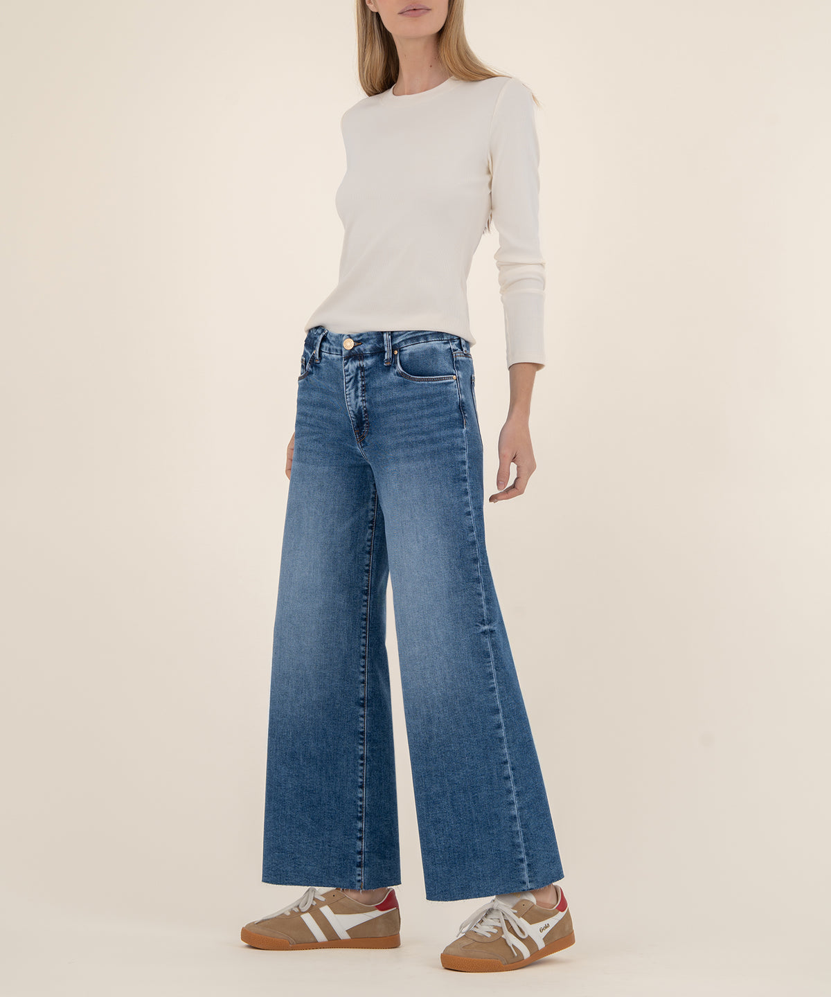 Woman wearing high-rise wide-leg blue jeans with a white long-sleeve top and tan sneakers.