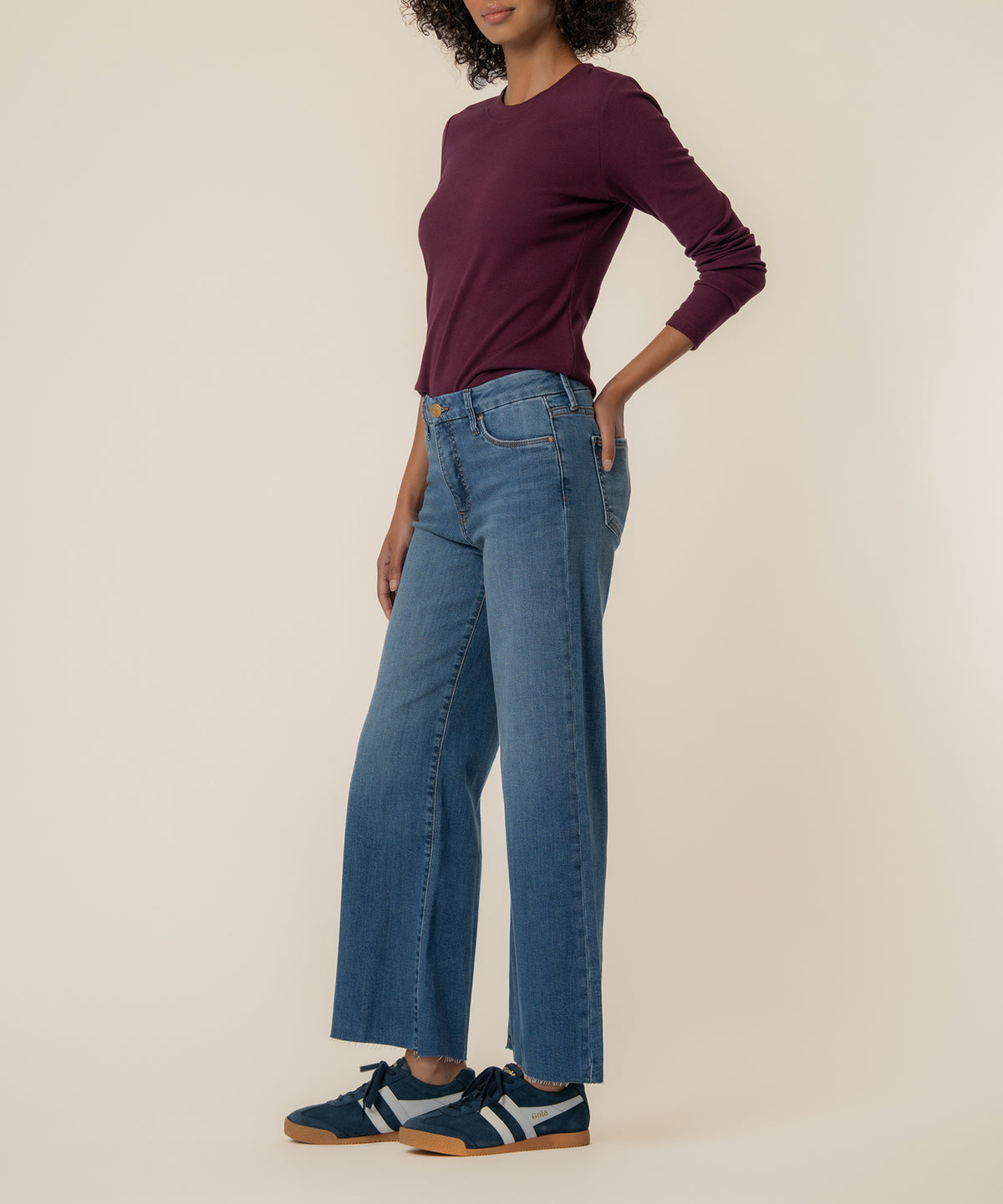 Woman wearing high-rise wide-leg jeans in light blue denim with purple long-sleeve top and navy sneakers.