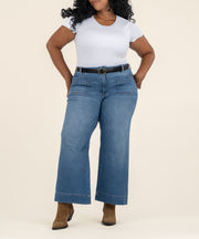 A woman wears high-rise medium-wash wide-leg jeans with front patch pockets, a white tee, black belt, and brown ankle boots.