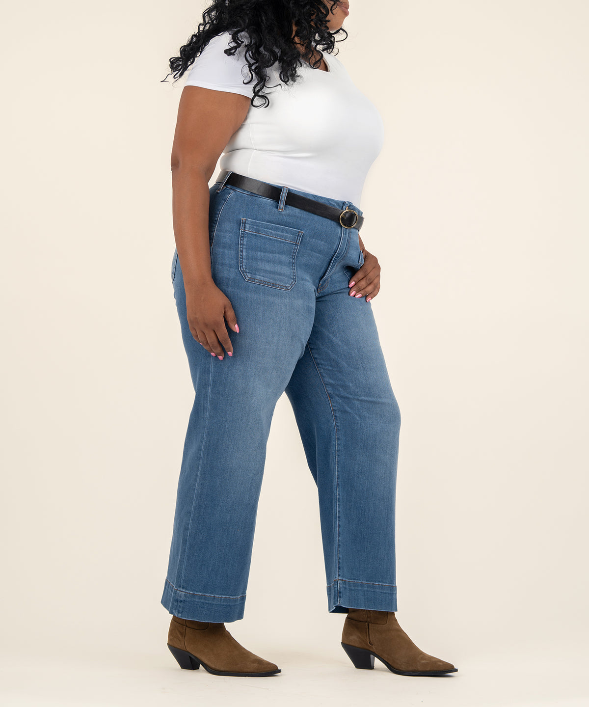 A model in side view wears medium-wash high-rise wide-leg jeans with a patch pocket, white top, black belt, and brown pointed ankle boots.