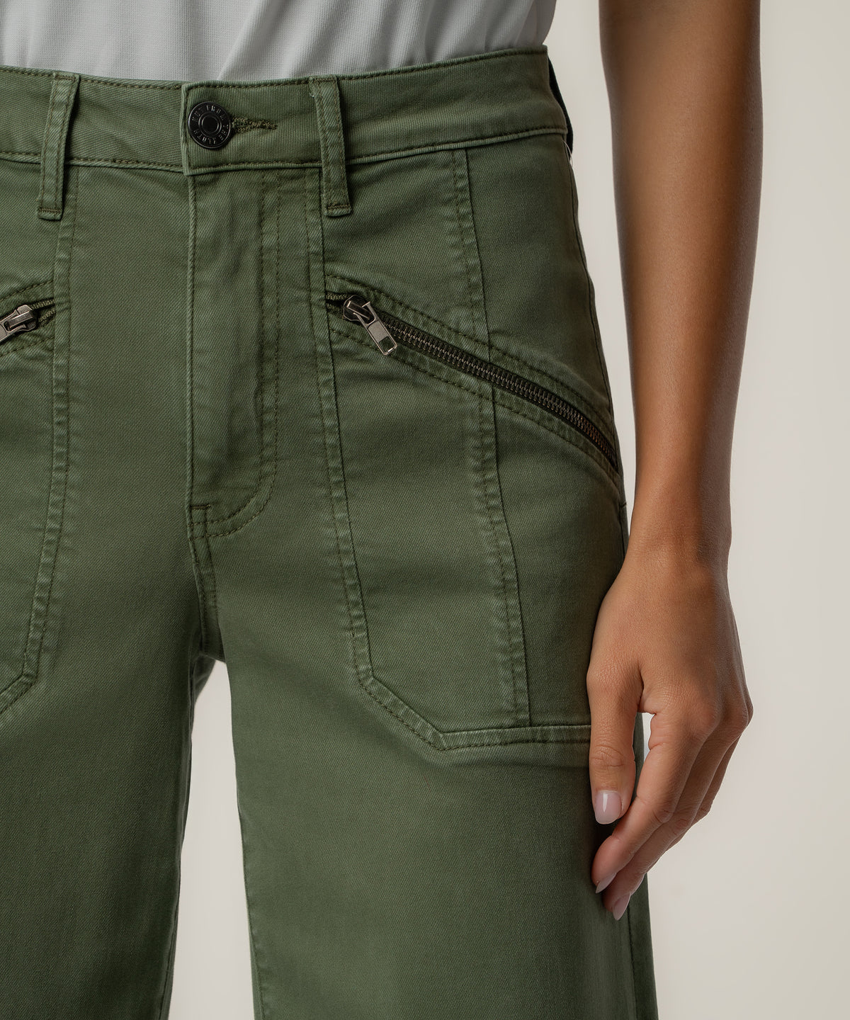 Close view of olive green high-rise wide-leg pants with angled zippered front pockets and a dark button, worn with a gray top.