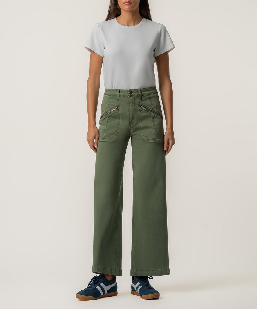 A person stands facing forward in olive green high-rise wide-leg pants with zippered front pockets, a light gray t-shirt, and navy sneakers.