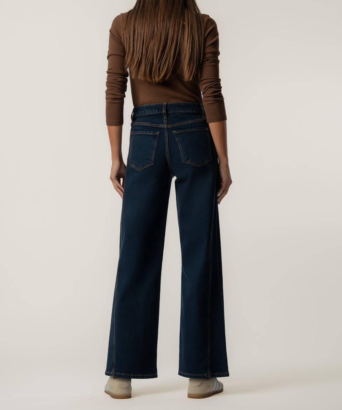 Rear view of a person wearing dark blue high-rise wide-leg jeans with a brown long-sleeve top and beige sneakers.