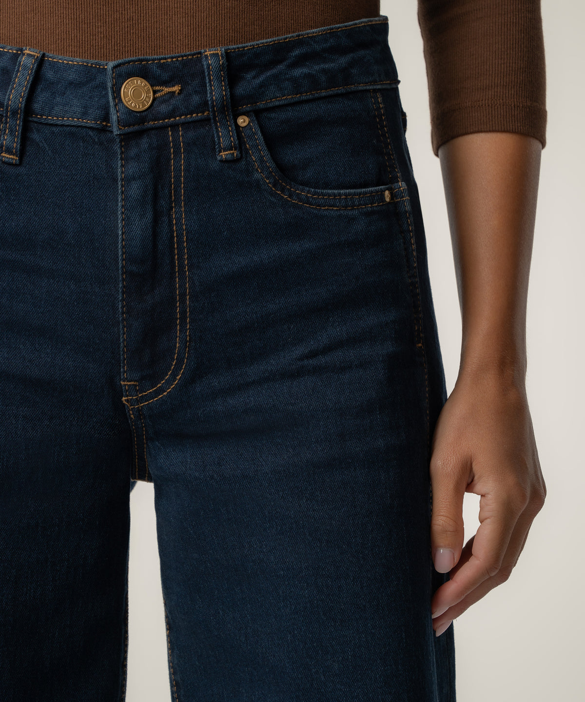 Close-up of dark indigo high-rise wide-leg jeans with gold button and orange stitching, worn with a brown ribbed top.