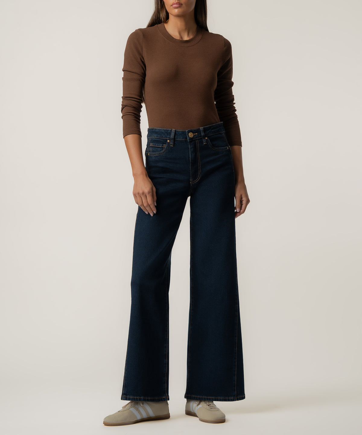 A woman stands against a plain background wearing dark indigo high-rise wide-leg jeans, a fitted brown long-sleeve top, and beige sneakers.