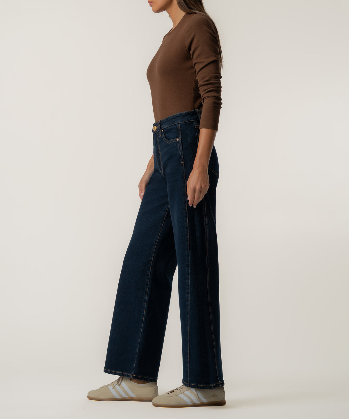 A woman in profile wears high-rise dark indigo wide-leg jeans with a brown long-sleeve top and beige sneakers.