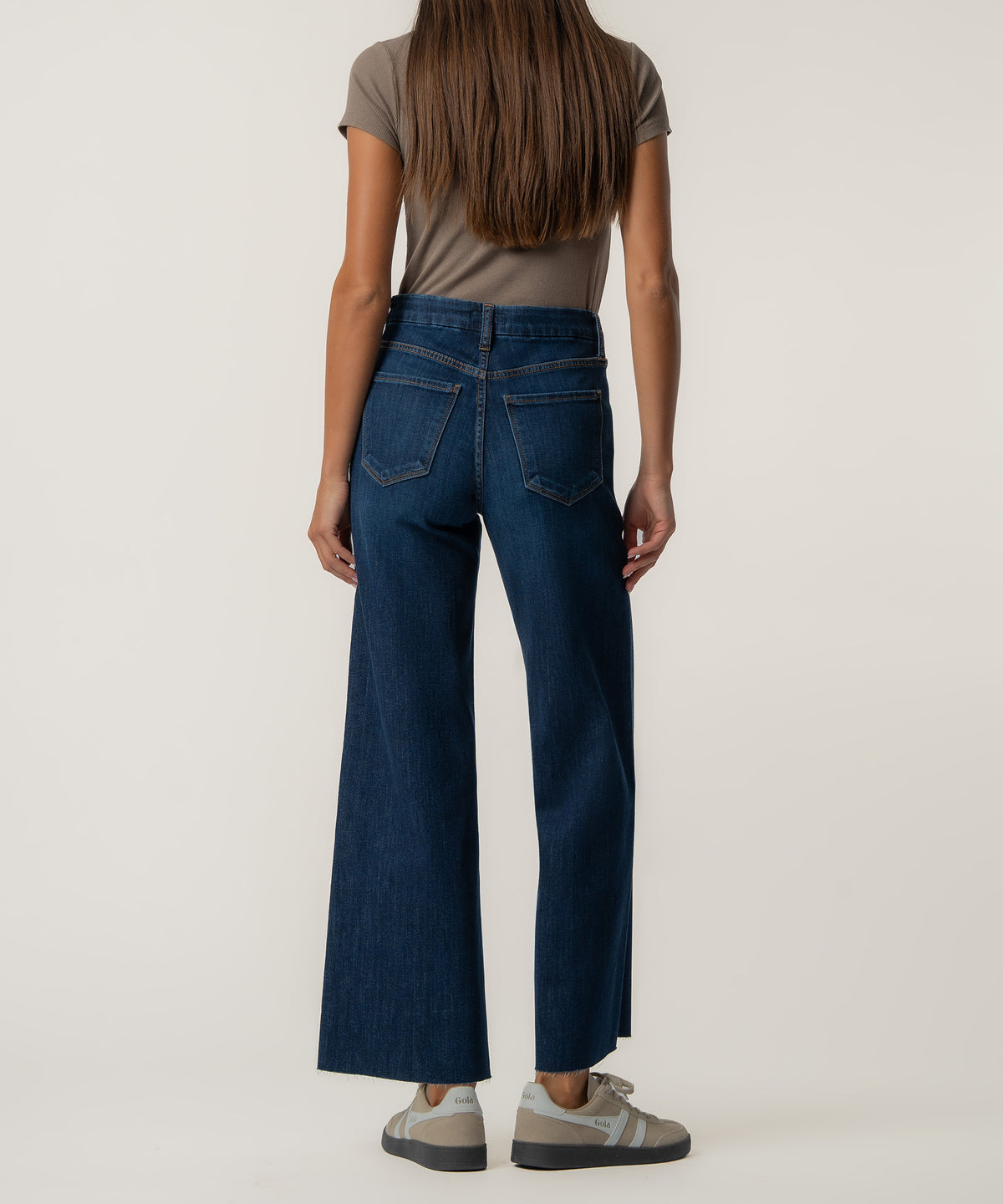 Back view of a woman wearing dark blue high-rise wide-leg jeans with raw hems, a taupe short-sleeve top, and beige sneakers.