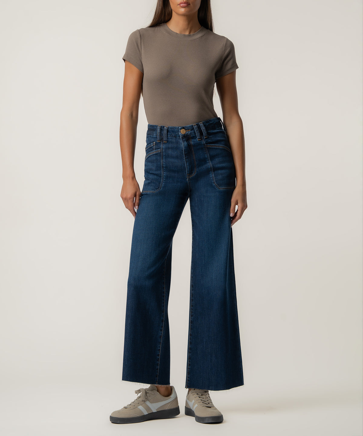 A woman stands wearing dark blue high-rise wide-leg jeans with front patch pockets, a taupe fitted t-shirt, and beige sneakers.