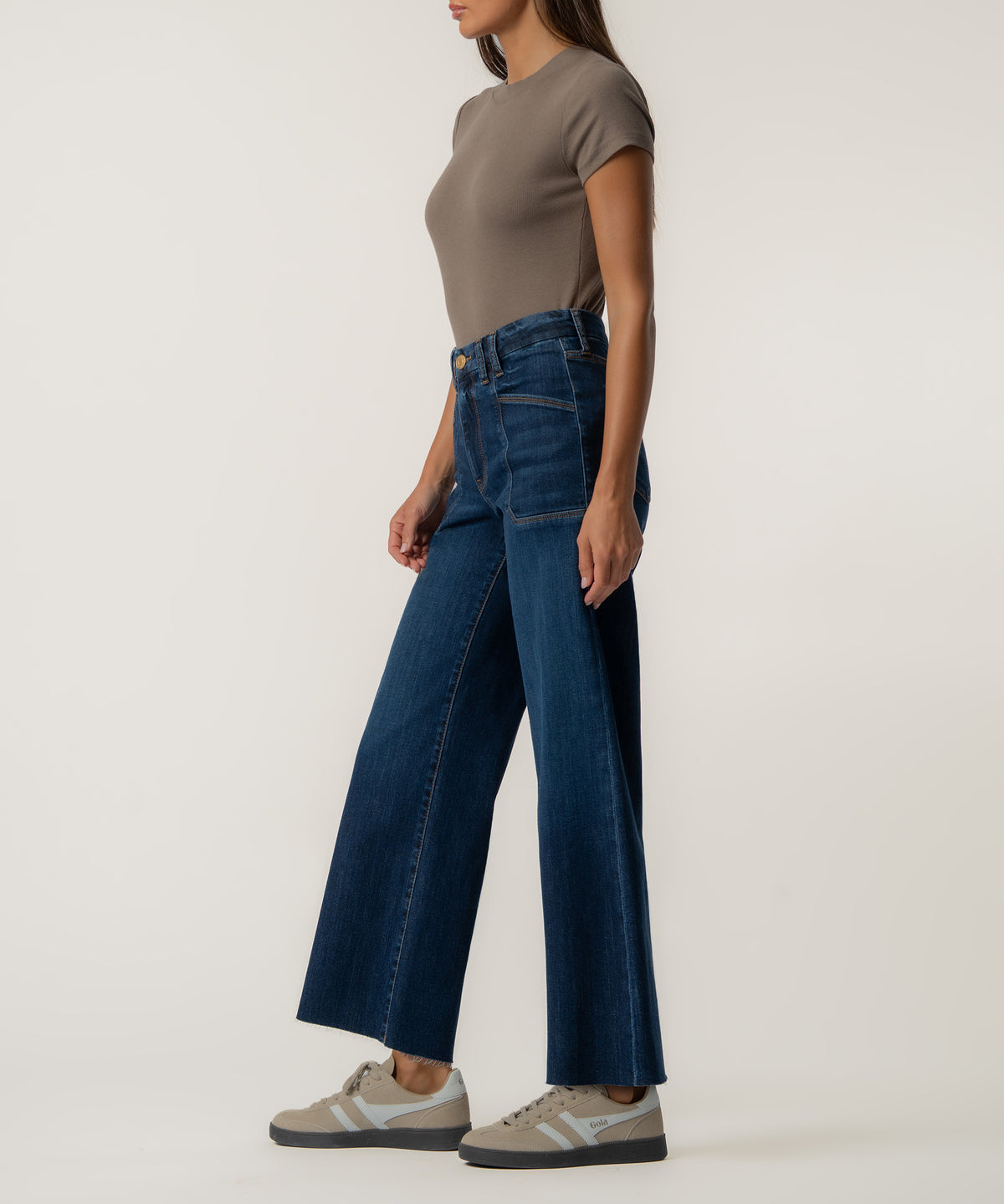 A woman stands in profile wearing high-rise dark blue wide-leg jeans with raw hems, a taupe short-sleeve top, and beige sneakers.