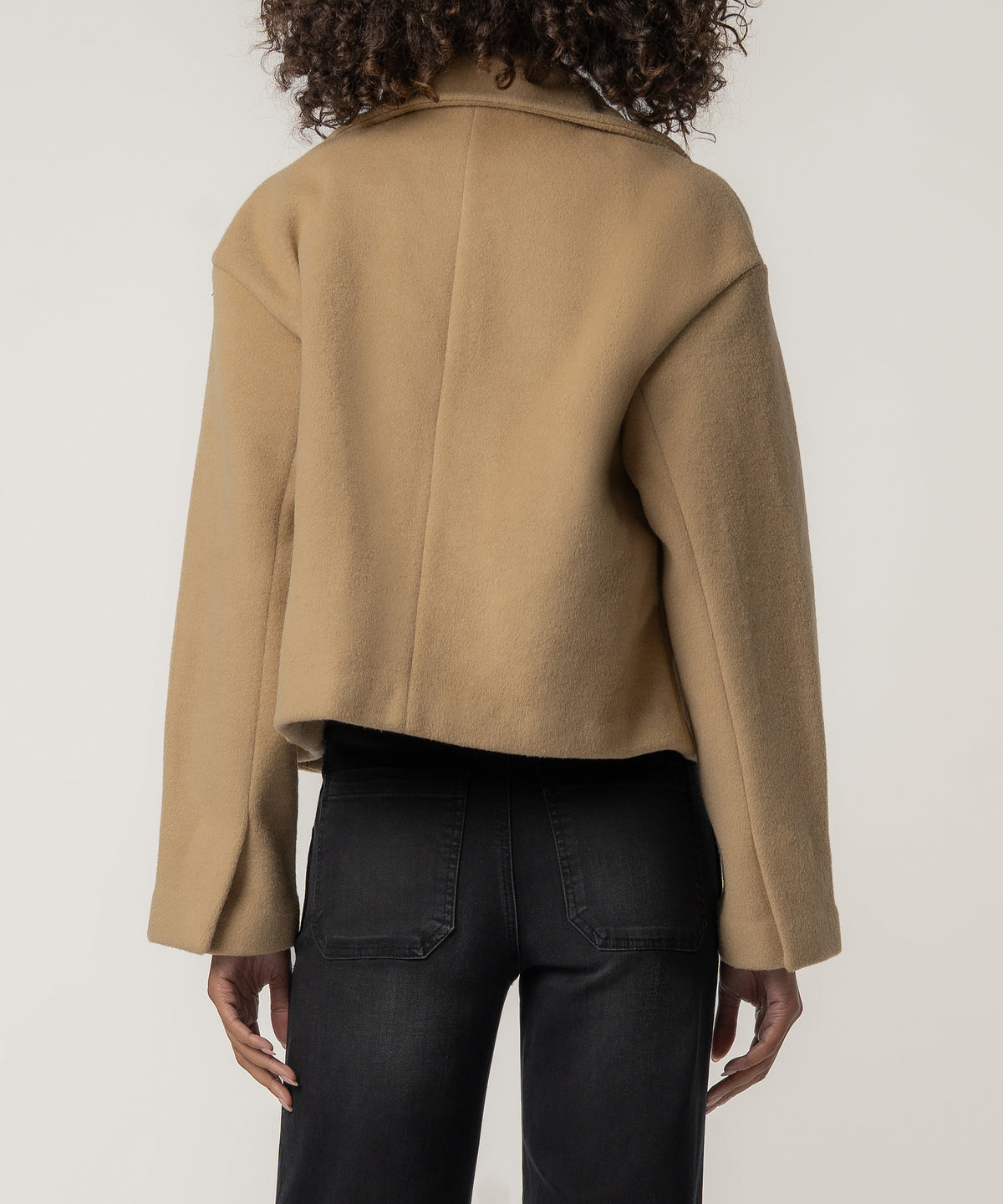 Back view of a person with curly hair wearing a cropped tan wool-style jacket with a collar and black jeans.