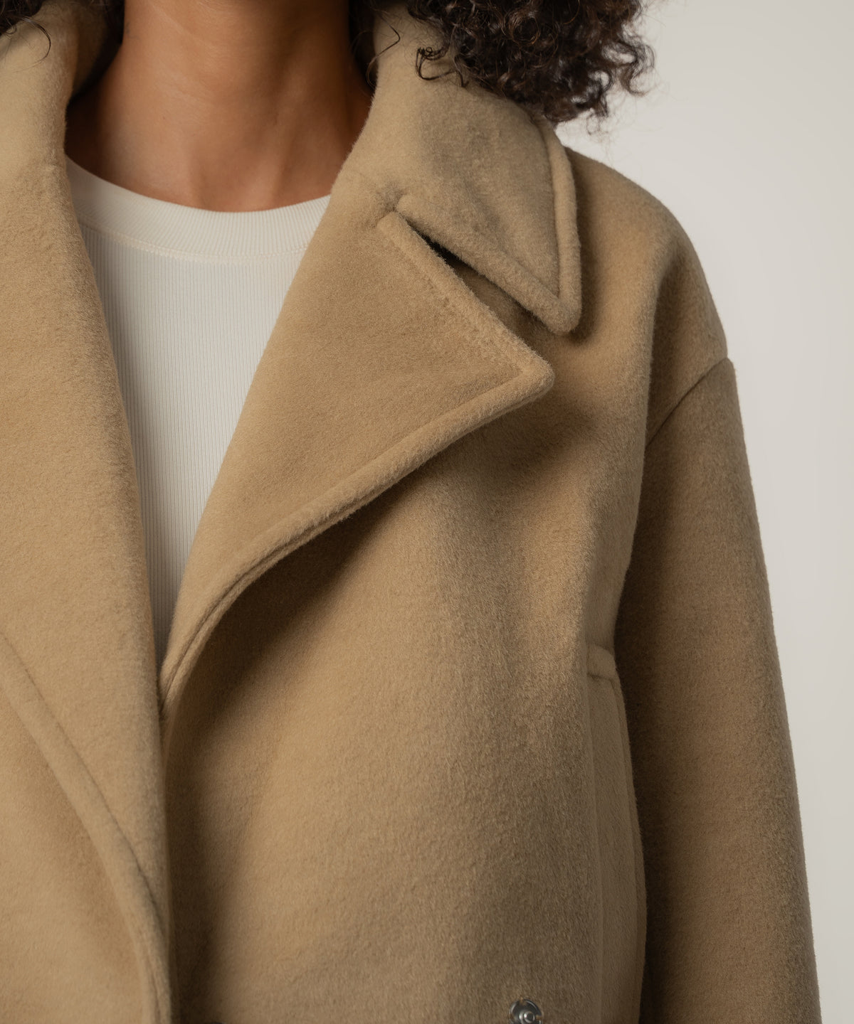 Close-up of a person with curly hair wearing a tan brushed wool jacket with wide lapels over a cream ribbed top.