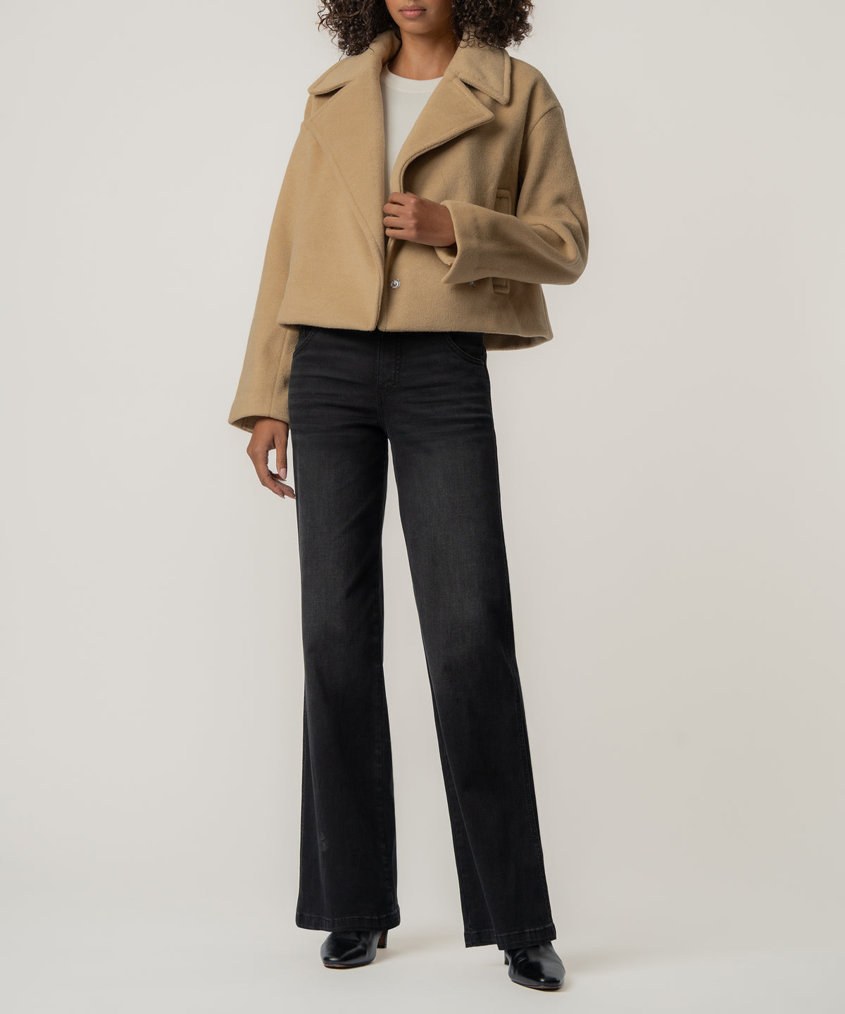 A woman wears a cropped tan wool-style jacket with wide lapels over a white top, black wide-leg jeans, and black boots.