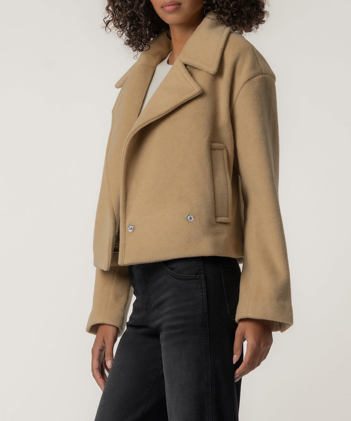 A woman wears a cropped tan wool jacket with wide lapels and snap closures, styled with a white top and black jeans.